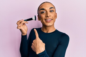 Hispanic man wearing make up and long hair holding makeup brush smiling happy pointing with hand...