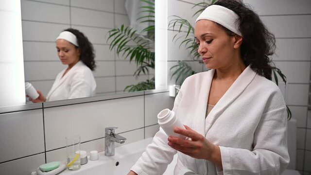 Confident middle-aged charming woman in white bathrobe opens a container with dietary supplement with vitamins and minerals, stands in front of the bathroom mirror, takes pills and drinks with water