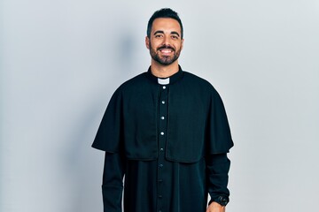 Handsome hispanic man with beard wearing catholic priest robe with a happy and cool smile on face....