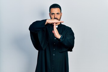 Handsome hispanic man with beard wearing catholic priest robe doing time out gesture with hands,...