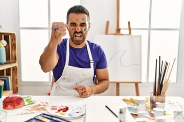 Young hispanic man with beard at art studio with painted face angry and mad raising fist frustrated and furious while shouting with anger. rage and aggressive concept.