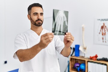 Young hispanic man wearing physiotherapist uniform holding hand xray at clinic