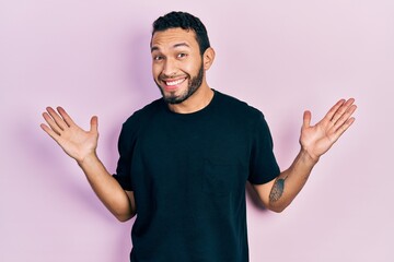 Fototapeta premium Hispanic man with beard wearing casual black t shirt clueless and confused with open arms, no idea and doubtful face.