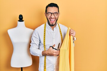 Hispanic man with beard dressmaker designer holding scissors and cloth smiling and laughing hard...