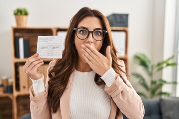 Young brunette woman holding covid record card covering mouth with hand, shocked and afraid for...