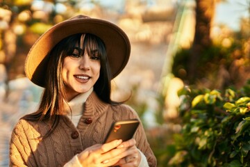 Brunette woman wearing winter hat smiling using smartphone at the park