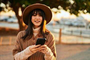 Brunette woman wearing winter hat smiling using smartphone outdoors at the city on sunset