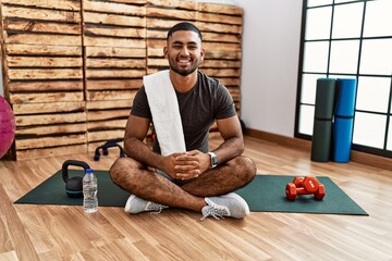 Young indian man sitting on training mat at the gym with a happy and cool smile on face. lucky person.