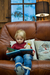 Blond toddler child, reading book and holding presents in a cozy cottage in the mountains