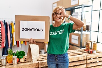 Middle age blonde woman wearing volunteer t shirt holding donations box smiling happy doing ok sign...