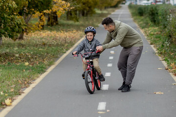 Obraz premium Father teaches his son to ride a bicycle on the bike path in the park. The father is holding a bicycle and the son is sitting on it