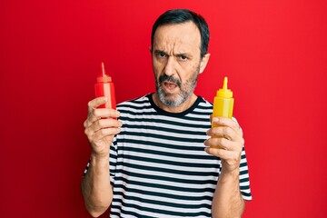 Middle age hispanic man holding ketchup and mustard bottle in shock face, looking skeptical and sarcastic, surprised with open mouth