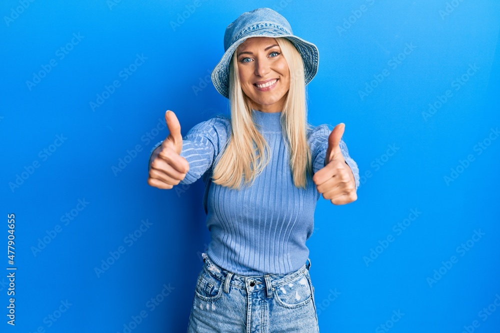 Wall mural Young blonde woman wearing casual denim hat approving doing positive gesture with hand, thumbs up smiling and happy for success. winner gesture.