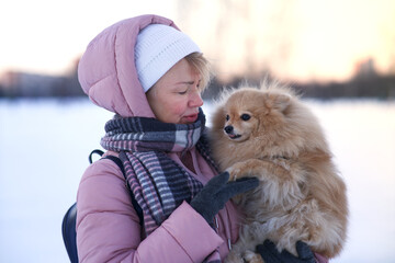 Happy beautiful woman, elderly senior mature lady is holding on hands, love, petting her adorable Pomeranian Spitz dog, cute puppy at winter snowy day in warm clothes hat, scarf outdoors in park