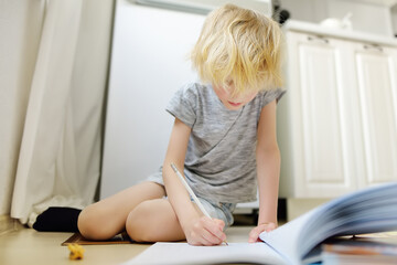 Elementary student boy doing homework on a floor at home. Child learning, doing exercises in workbook. Math and language tutorial. Preparing preschooler baby for school.