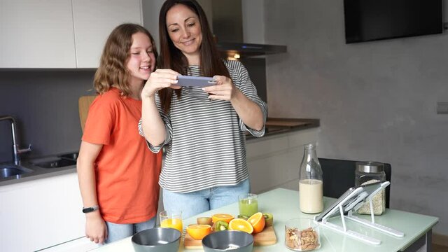 Happy mother and teen daughter take photo with mobile to their healthy breakfast of fruit juice, cereals and oatmeal drink. Concept of loving and caring for oneself and spending time with the family.
