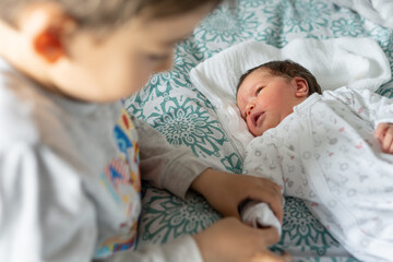 Close up on newborn caucasian baby lying in bed on first day of her life with her little brother beside holding her hand and taking care of him - brotherhood and growing up family bonding concept