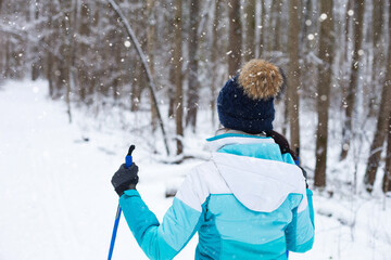 Skier in windbreaker and hat with pompom with ski poles in his hands with his back against the background of a snowy forest. Cross-country skiing in winter forest, outdoor sports, healthy lifestyle.