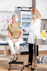 A Young White Female Medical Doctor Administering a Covid-19 Vaccine with a Syringe Needle to an Elderly Senior Female Patient Wearing Generic ID Badge, Gloves and Mask in Hospital or Clinic.