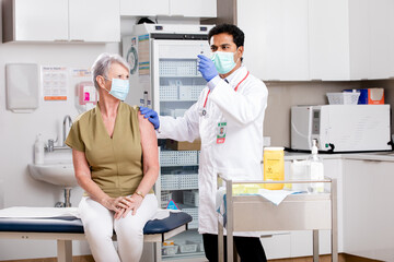 A Young Indian Male Medical Doctor Administering a Covid-19 Vaccine with a Syringe Needle to an Elderly Senior Female Patient Wearing Generic ID Badge, Gloves and Mask in Hospital or Health Clinic.