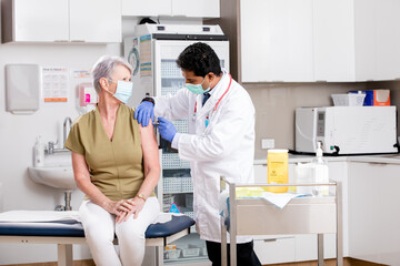 A Young Indian Male Medical Doctor Administering a Covid-19 Vaccine with a Syringe Needle to an Elderly Senior Female Patient Wearing Generic ID Badge, Gloves and Mask in Hospital or Health Clinic.