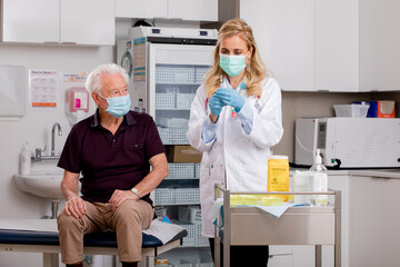 A Young White Female Medical Doctor Administering a Covid-19 Vaccine with a Syringe Needle to an Elderly Senior Male Patient Wearing Generic ID Badge, Gloves and Mask in Hospital or Health Clinic.