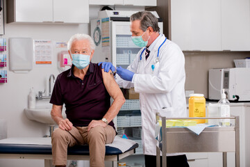 A White Male Medical Doctor Administering a Covid-19 Vaccine Injection with a Syringe Needle to an Elderly Senior Male Patient Wearing Generic ID Badge, Gloves and Mask in Hospital or Health Clinic.