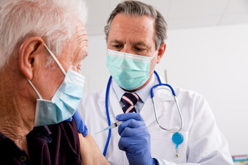 Fototapeta na wymiar A White Male Medical Doctor Administering a Covid-19 Vaccine Injection with a Syringe Needle to an Elderly Senior Male Patient Wearing Generic ID Badge, Gloves and Mask in Hospital or Health Clinic.
