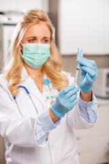 A Young Female Medical Doctor Extracting a Covid-19 Vaccine with a Syringe Needle Wearing Generic ID Badge, Gloves, Mask, White Lab Coat and Stethoscope in Hospital or Health Clinic.