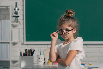 Close-up portrait of attractive small little cheerful girl sitting on table desktop in class room indoors. Little funny school girl face. Nerd school girl in glasses on chalkboard.