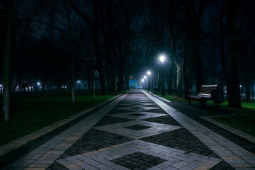 The alley of a night early winter park in a light fog. Footpath in a fabulous late autumn city park at night with benches and latterns. Beautiful cold evening in Mariinsky Park. Kyiv, Ukraine.