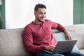 Cheerful arab guy sitting on couch at home, using laptop