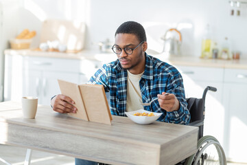Millennial handicapped black man in wheelchair eating breakfast and reading book indoors