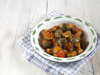 Sambal balado terong bulat or stir fried brinjal or round eggplant with chili sauce. Typical hot and spicy Indonesian traditional dish. Served in white plate on white wooden table.