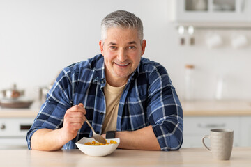 Fototapeta na wymiar Healthy breakfast concept. Cheerful senior man having breakfast, eating cereals and drinking coffee, free space
