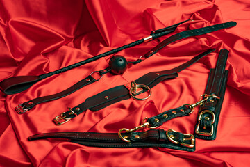Adult sex games. BDSM items. Leather straps handcuffs, collar, gag ball and whip on a red satin sheet.