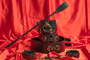 Adult sex games. BDSM items. Leather straps handcuffs, collar, belt and whip on a red satin sheet.