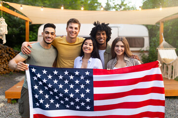 Diverse millennial friends holding American flag and smiling at camera in front of motorhome at...