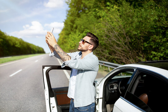 Unhappy Caucasian man standing near car, having problem with mobile connection, trying to find network on highway