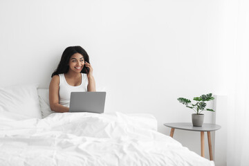 Multitasking. Happy black woman talking on smartphone and using laptop computer, sitting on bed at home, empty space