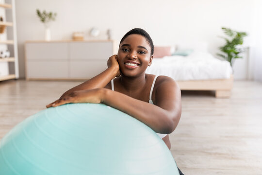 Happy Plus Size Black Woman Sitting Near Fitness Ball, Smiling At Camera Indoors