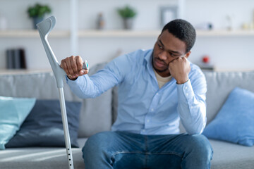 Unhappy African American guy leaning on crutch, suffering from depression after injury at home, selective focus