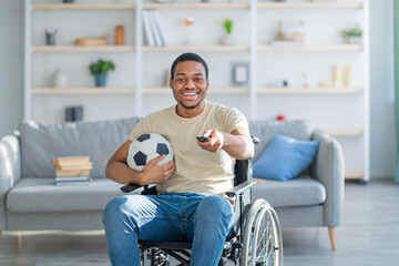 Cheerful impaired black guy in wheelchair watching football game on TV at home