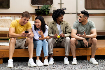 Diverse friends sitting near motorhome, talking to each other, using smartphone, spending time together on camping trip
