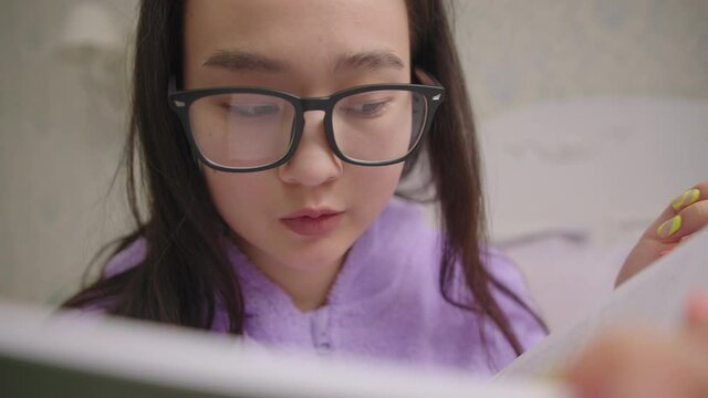 Close up of Asian woman in eyeglasses reading book sitting in bed. Handheld camera, slow motion.
