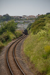 Fototapeta na wymiar Steam train on tracks between green hills and colourful houses in the background