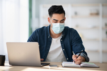 Arab man in protective face mask using laptop, taking notes