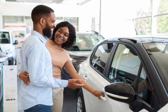 Car Choosing. Happy Black Spouses Standing Next To Modern Vehicle In Showroom