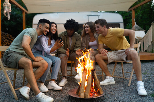 Group Of Cheerful Diverse Friends Spending Time Near Bonfire, Having Camping Vacation With RV, Having Fun Outdoors