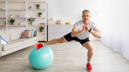 Happy sporty mature man doing side lunges, using fitness ball, working out in living room interior, panorama, copy space